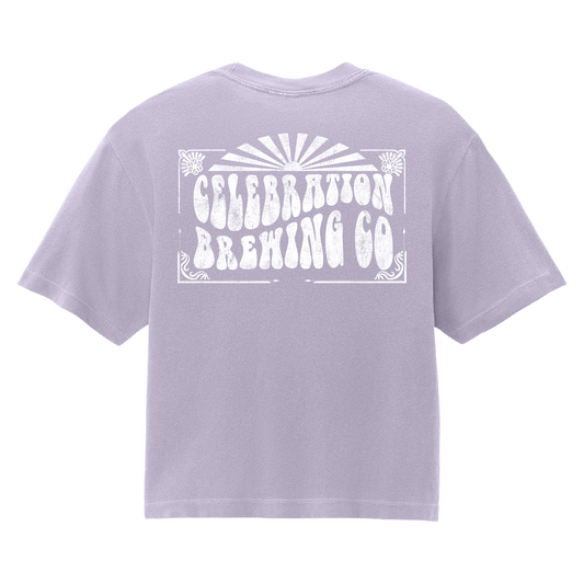 A light lavender purple short-sleeve t-shirt shown from the back, featuring a large white distressed graphic that reads 'CELEBRATION BREWING CO' with a sunburst design above it.