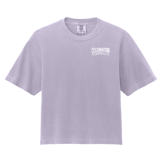 A light lavender crewneck t-shirt with a white graphic print on the left chest reading 'CELEBRATION' and 'ONE CELEBRATING EVERY DAY'. The shirt is displayed flat against a solid white background.